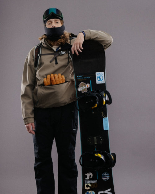 Person in winter gear with a snowboard, holding sausages in a vest pocket.