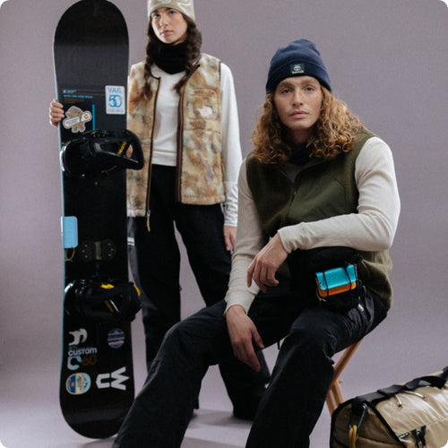 Two people in winter clothing, one seated with a snowboard, against a gray background. Both wear beanies and layered outfits.