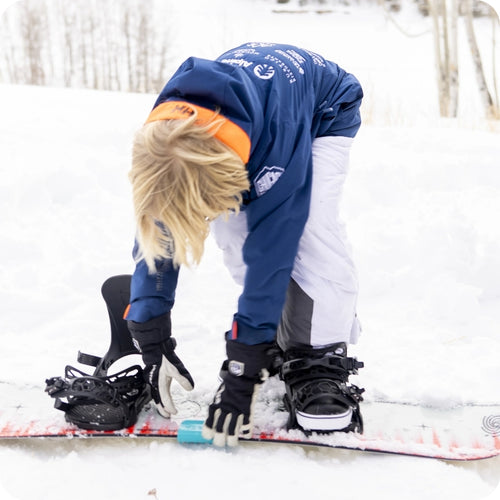 A person kneels on snow, adjusting a snowboard with a blue tool, wearing a blue jacket and white pants.