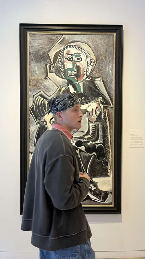 A person stands in front of an abstract painting in a gallery, wearing a headscarf, sweatshirt, and jeans.