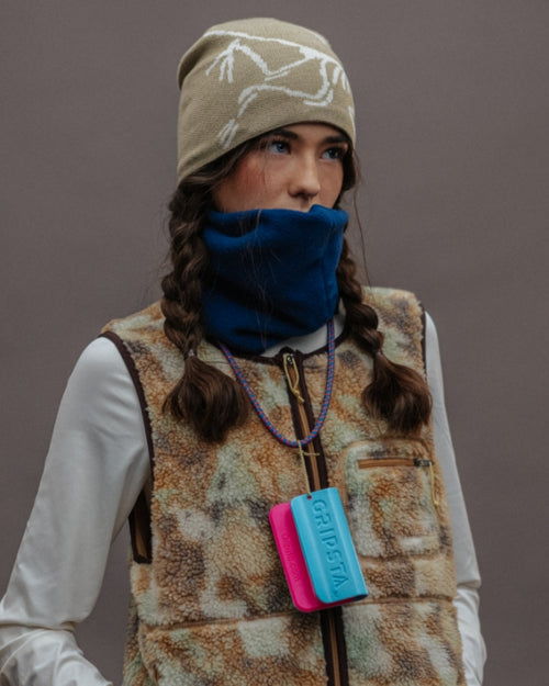 Person wearing a beanie and vest with a blue neck warmer, holding a colorful device, stands against a gray background.