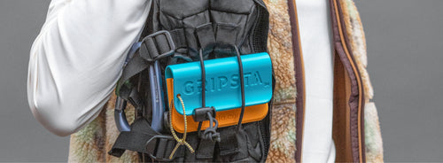 Person carrying a backpack with a blue and orange GRIPSTÅ case attached, wearing a brown and beige vest over a white shirt.