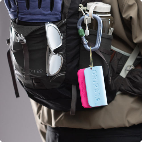 A close-up of a backpack with sunglasses, gloves, a carabiner, a water bottle, and a blue and pink GRIPSTA attached.