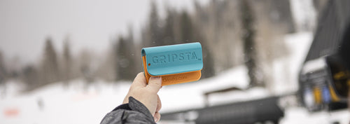 Hand holding a blue and orange GRIPSTIC, with snowy trees and blurred background.