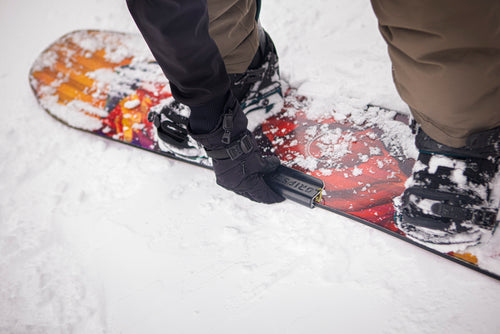 A person in gloves adjusting bindings on a colorful snowboard in the snow.