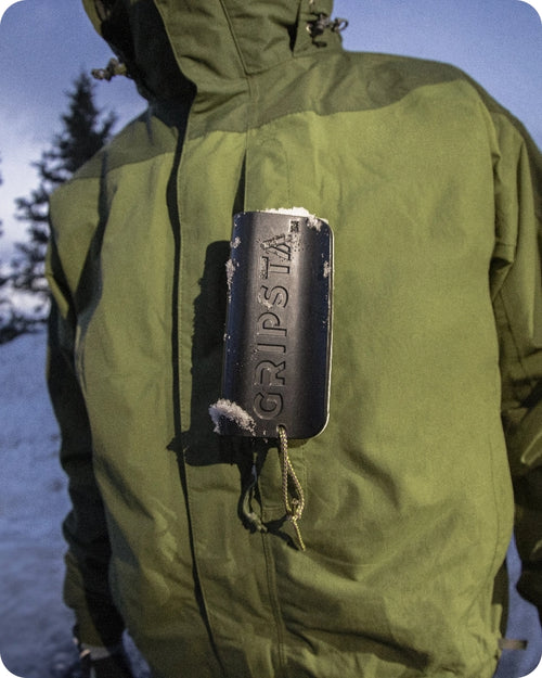 Person in a green jacket outdoors, holding a black Gripsta tool covered in snow against their chest.