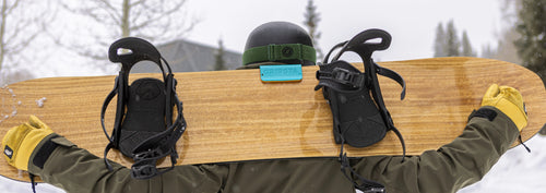 A person wearing a helmet and gloves carries a snowboard on their shoulders in a snowy landscape.