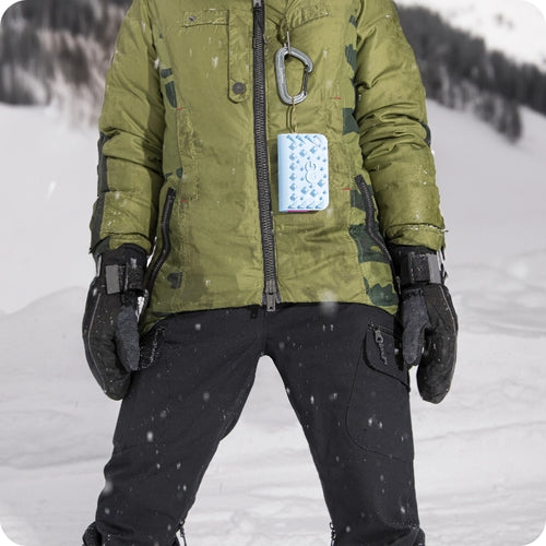 Person in a green jacket and black gloves standing in the snow, with a blue ice pack attached to the jacket.