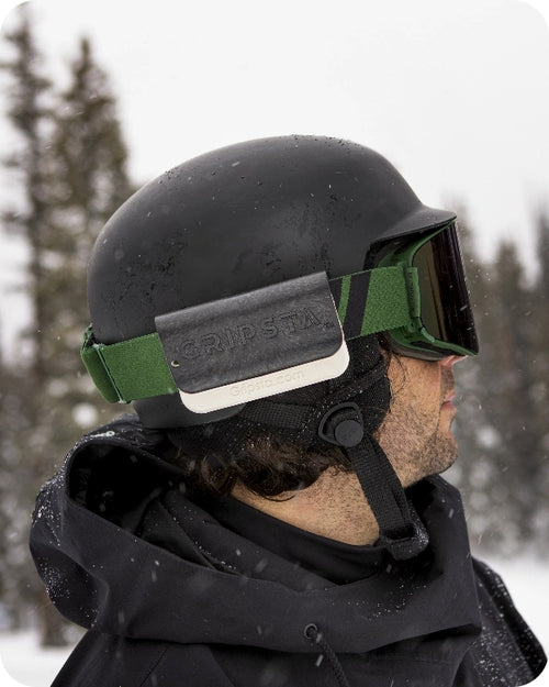 A person wearing a black ski helmet and goggles stands in the snow with trees in the background.