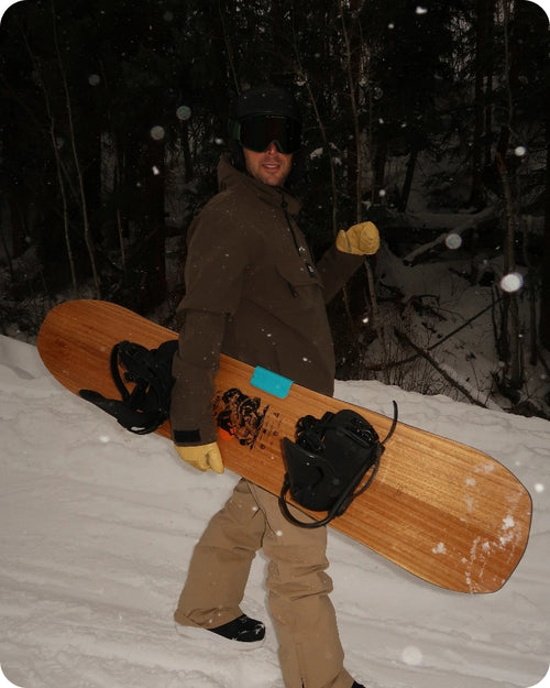 A person in snow gear holding a snowboard, standing on a snowy path in a forested area.