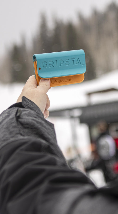 A person holds a blue and orange Gripsta device, with a snowy outdoor scene in the background.